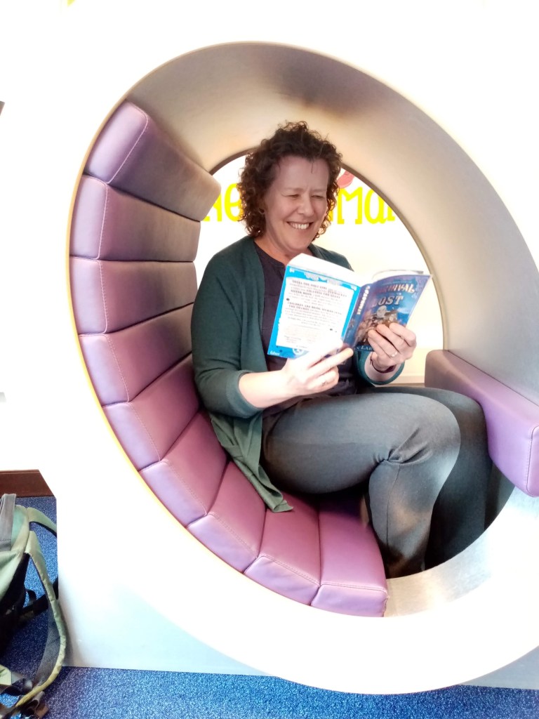 Sandra sits in a round children's seat with purple padding. She is reading a children's book and smiling