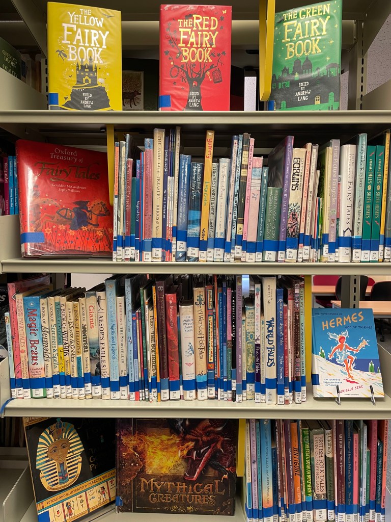 Four library shelves containing children's books. On each shelf, there is one or more books facing outwards, including books on mythical creatures and fairies.