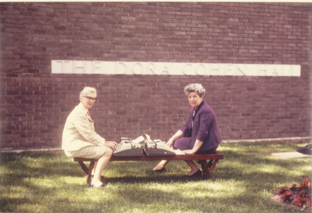 Two women sit outdoors on a wooden bench. Between them are two typewriters. Behind them is a brick wall with a plaque reading The Dora Cohen Hall.