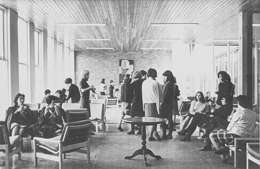 A room is filled with casual chairs and tables. Around twenty female students are mingling in the space.