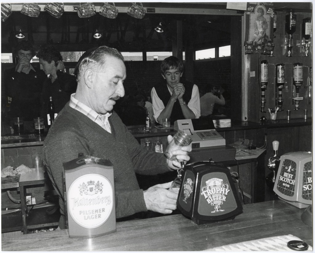 A member of bar staff pulls a pint of bitter. Behind him, a couple of students are leaning on the bar.