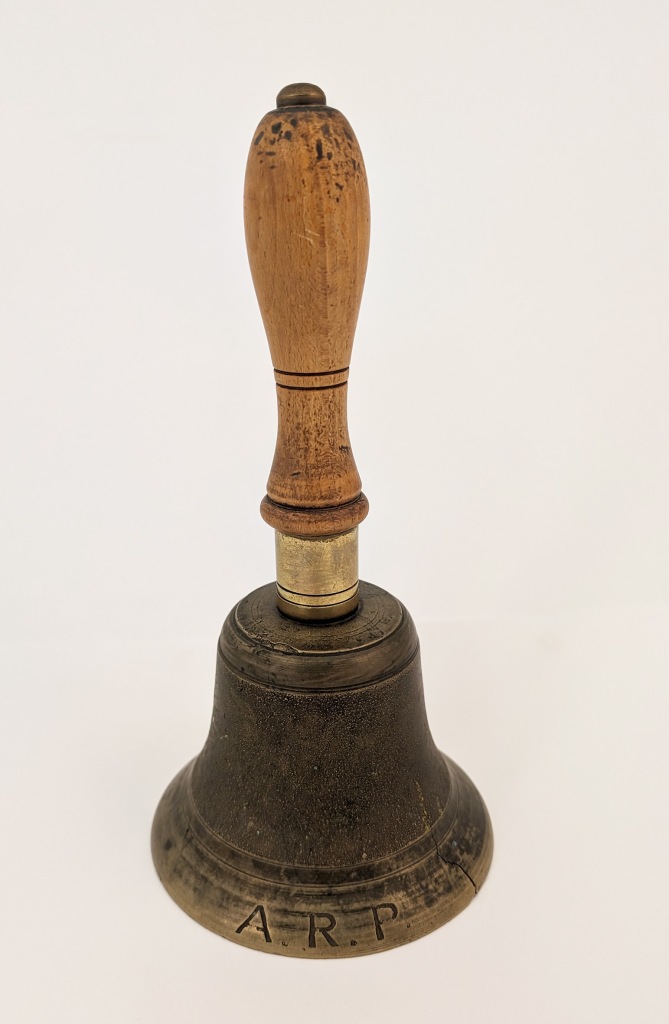 A brass handbell with a wooden handle. The brass is engraved with the initials ARP.