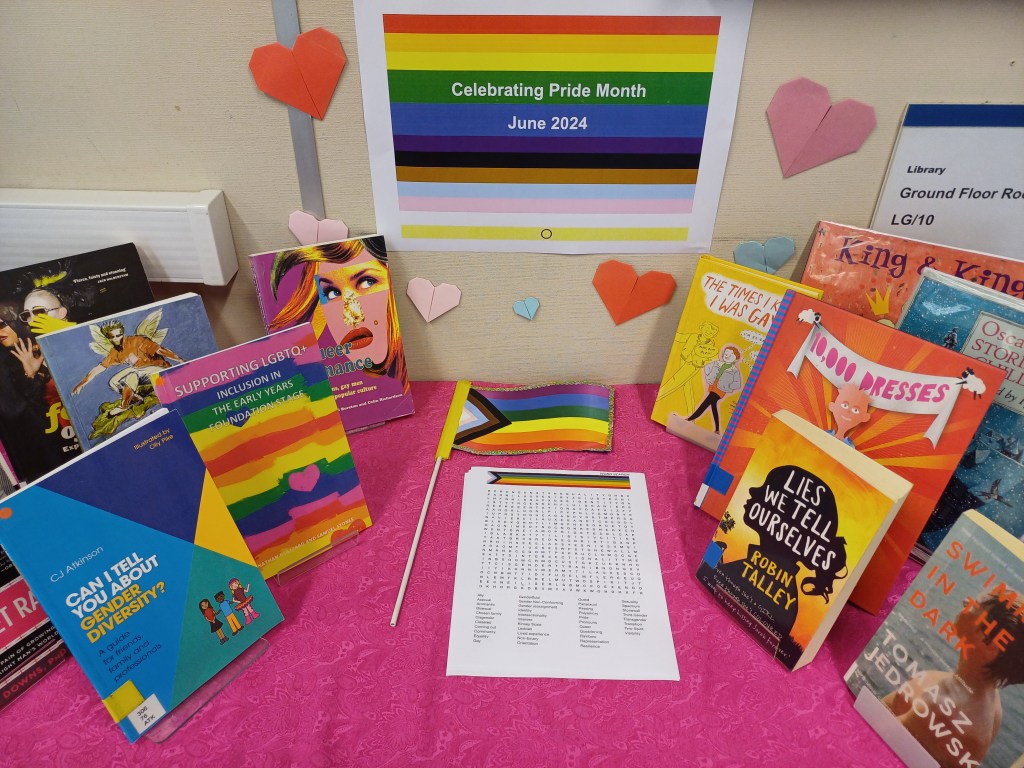 A table contains a pride flag, a wordsearch and six books. Stuck on the wall above is a pride sign and some origami paper hearts.